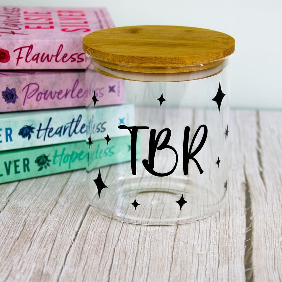 Glass jar with bamboo lid lettered “TBR” with stars, in front of a stack of books