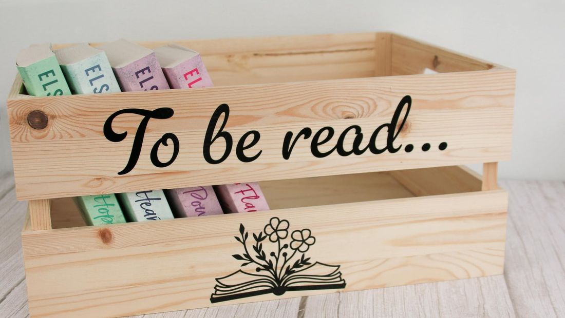 Wooden crate lettered “To be read…” with an open book and flowers, holding a row of books
